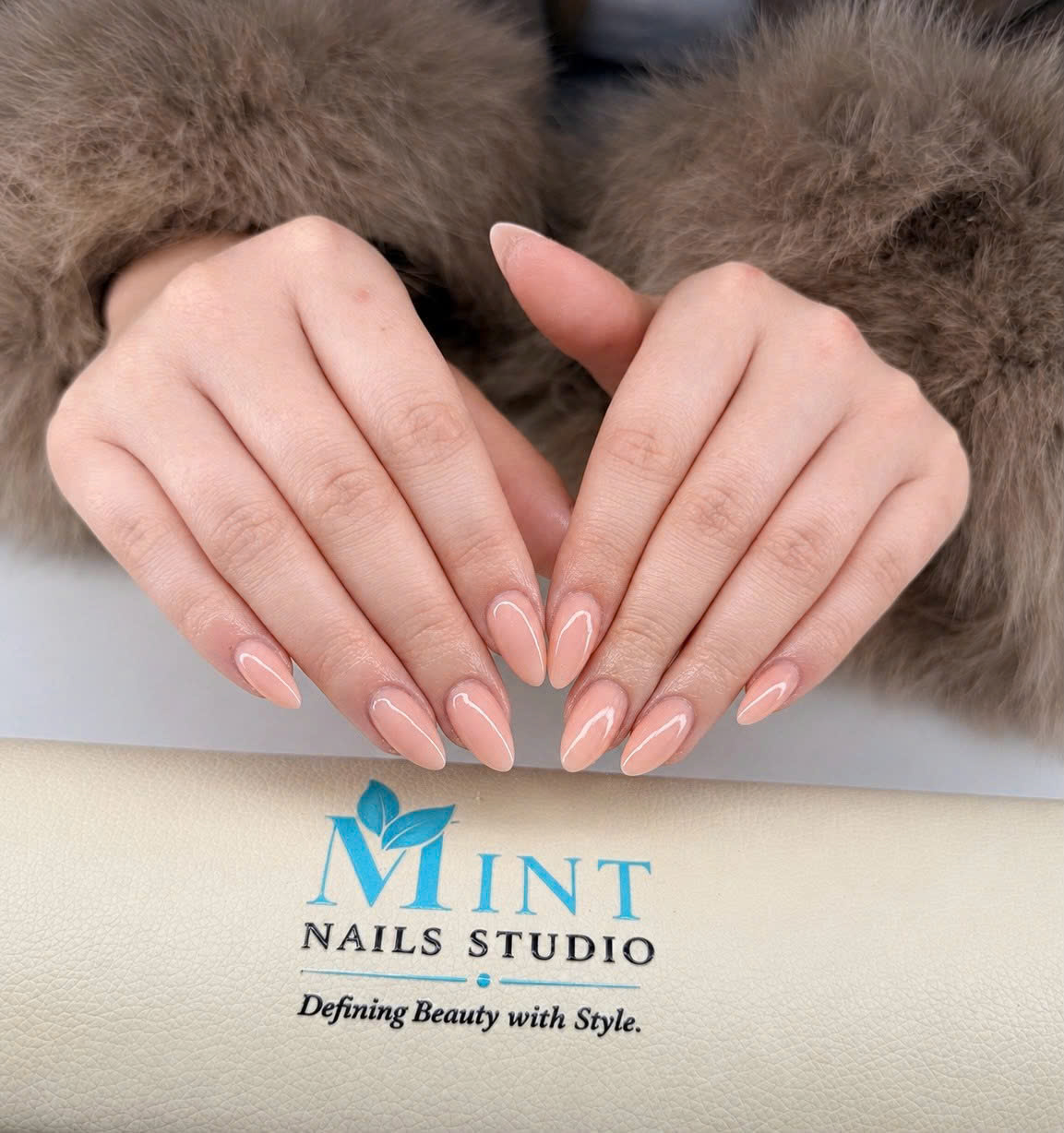 Glänzendes Nude-Nageldesign in Mandelform vor dem Mint Nails Studio Logo